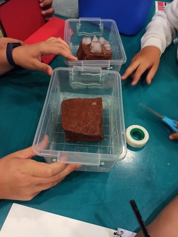 Two clear plastic boxes on a table, each holding a brick, with ice cubes in one of them. Children's hands hold the boxes.