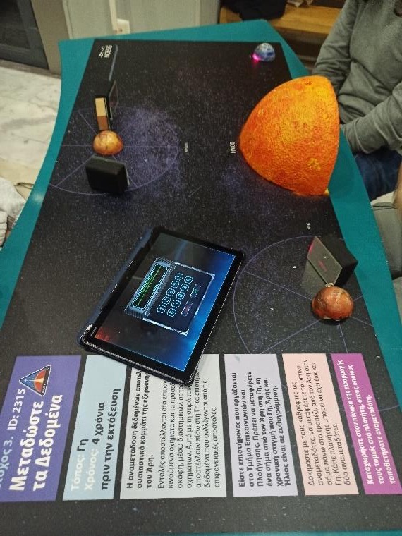 A dark game board with model planets, a large orange half-sphere for the Sun, a tablet showing a keypad, and task cards in Greek along the edge.