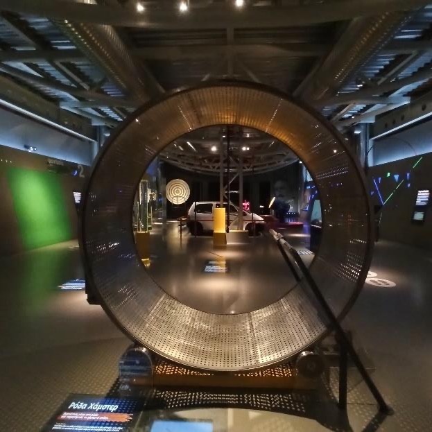 A large metal wheel, big enough for a person to walk inside, standing in a dim exhibition hall.