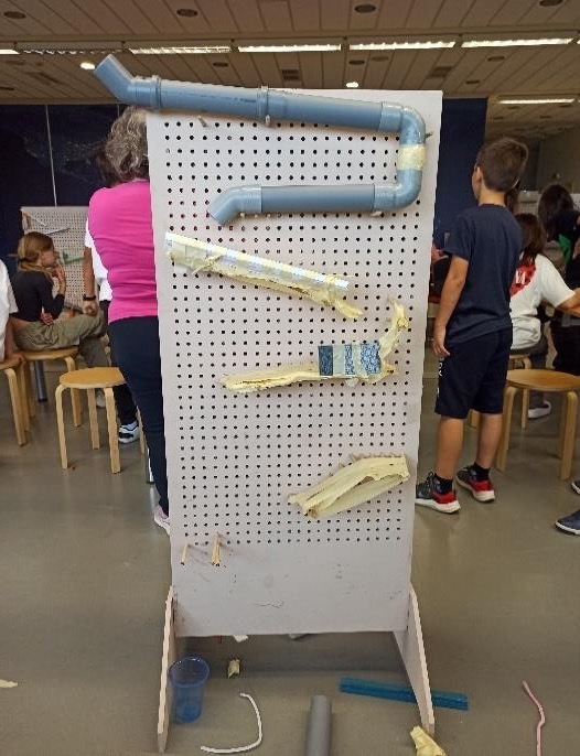 A white pegboard wall with pipes, cardboard ramps and tape attached, with pupils working around it.